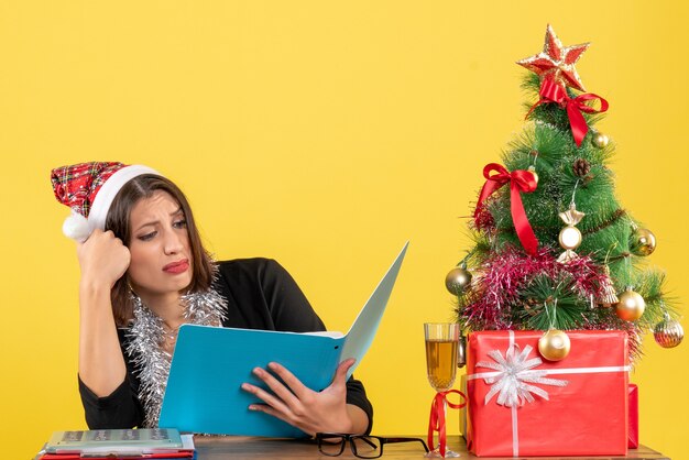 Mulher de negócios de terno com chapéu de papai noel e decorações de ano novo focada no documento e sentada em uma mesa com uma árvore de natal no escritório