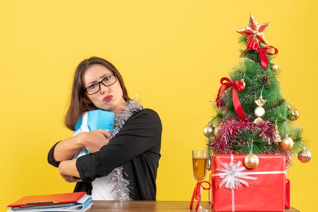 Mulher de negócios de terno com óculos escondendo seu presente de forma surpreendente e sentada em uma mesa com uma árvore de natal no escritório