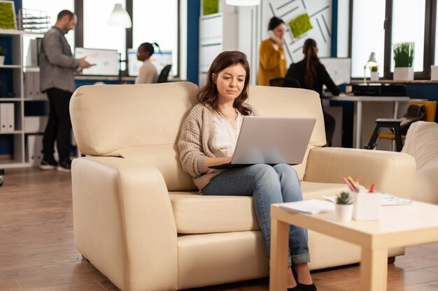 Mulher de negócios digitando em um laptop, sentada no sofá do escritório inicial, enquanto uma equipe diversificada trabalha em segundo plano, analisando dados estatísticos