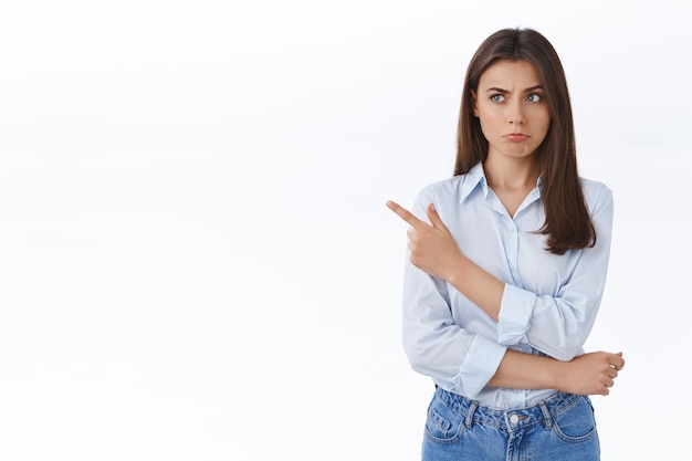 Foto grátis mulher de negócios duvidosa e insegura parecendo cética com leve descrença, fazendo beicinho pensativo enquanto aponta o dedo esquerdo para uma oferta estranhamente boa, hesitando e ponderando sobre a parede branca