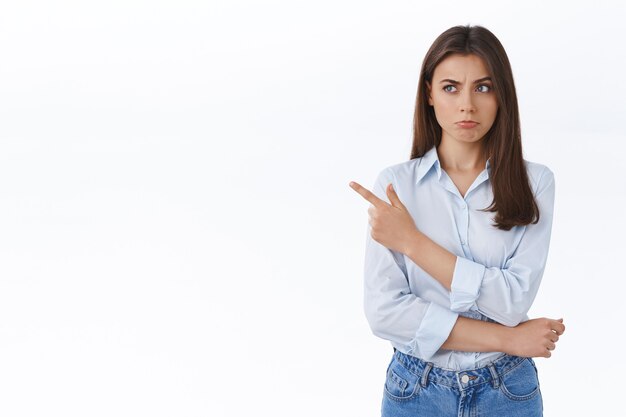 Mulher de negócios duvidosa e insegura parecendo cética com leve descrença, fazendo beicinho pensativo enquanto aponta o dedo esquerdo para uma oferta estranhamente boa, hesitando e ponderando sobre a parede branca