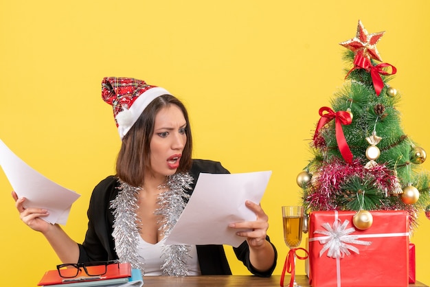 Mulher de negócios emocional de terno com chapéu de Papai Noel e decorações de ano novo, verificando documentos e sentada em uma mesa com uma árvore de natal no escritório