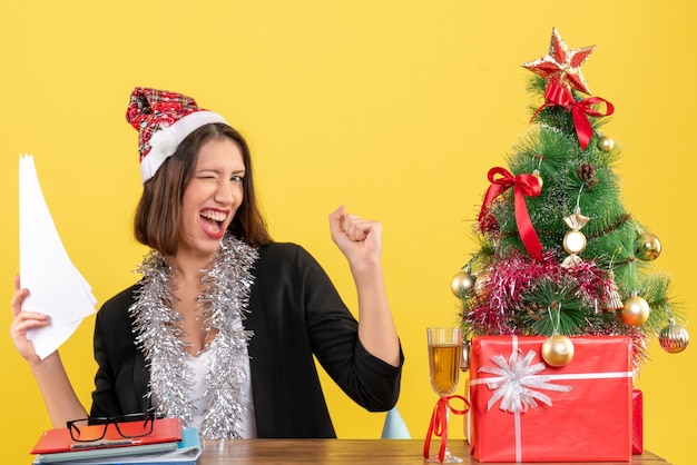 Mulher de negócios emocional em um terno com chapéu de Papai Noel e decorações de ano novo segurando documentos e sentada em uma mesa com uma árvore de natal no escritório