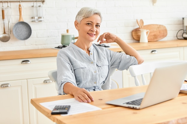 Mulher de negócios energética moderna de idade madura, sentada à mesa de jantar, tomando café da manhã e verificando e-mail usando um computador portátil. Freelancer feminina sênior elegante trabalhando em casa em um laptop