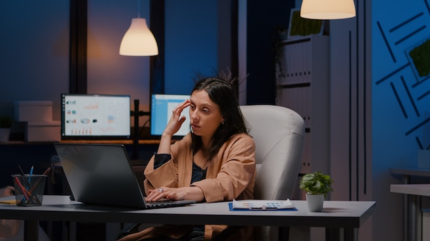 Mulher de negócios exausta e sobrecarregada trabalhando no escritório de inicialização, verificando a estratégia de gerenciamento no laptop tarde da noite