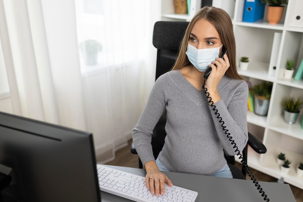 Mulher de negócios grávida falando ao telefone enquanto está na mesa e usando máscara médica