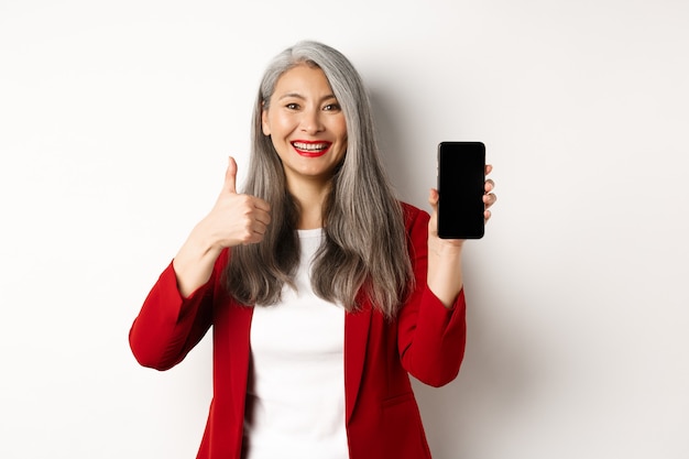 Mulher de negócios idosa asiática satisfeita mostrando a tela do smartphone em branco e o polegar para cima, elogiando a promoção online ou o aplicativo da empresa, em pé sobre um fundo branco