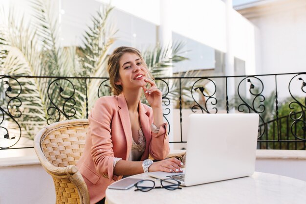 Mulher de negócios jovem bonita, estudante sentada no terraço, no café, trabalhando no laptop, aproveitando o trabalho. Vestindo uma elegante jaqueta rosa, relógios brancos. Óculos e smartphone na mesa.