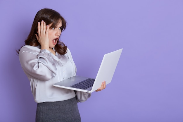 Mulher de negócios jovem nervosa atraente com o laptop nas mãos, olhando para a tela do dispositivo com expressão chocada, tocando sua cabeça com a mão, mantém a boca bem aberta, fica contra a parede lilás.