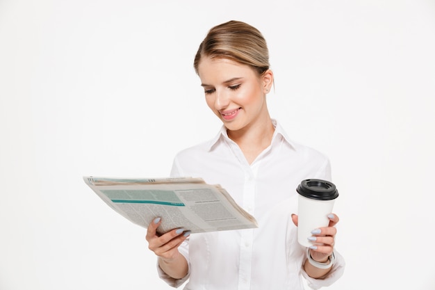 Foto grátis mulher de negócios loira alegre lendo jornal enquanto segura a xícara de café sobre parede branca