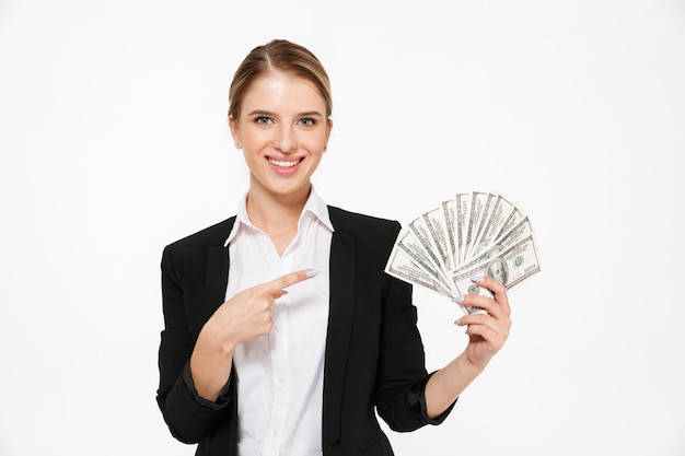 Mulher de negócios loira sorridente segurando o dinheiro e apontando o seu tempo sobre parede branca