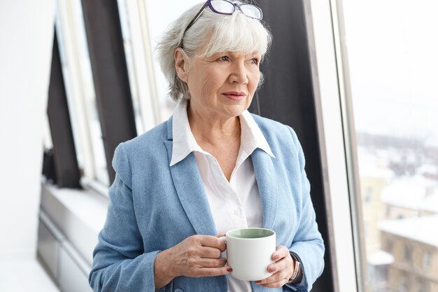 Mulher de negócios madura de cabelos grisalhos séria usando óculos na cabeça e elegantes roupas formais, desfrutando de um café quente, em pé na janela com a xícara nas mãos, com um olhar pensativo e pensativo
