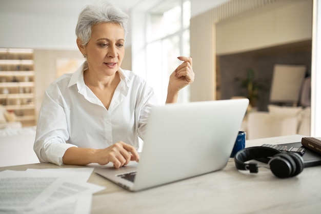 Mulher de negócios madura elegante com corte de cabelo curto sentada em frente ao laptop, olhando para a tela com a boca aberta como se estivesse dizendo algo