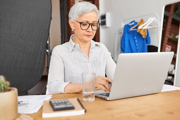 Mulher de negócios madura qualificada experiente confiante usando óculos elegantes, escrevendo cartas para seus clientes e parceiros de negócios, usando a conexão de internet sem fio de alta velocidade no laptop.