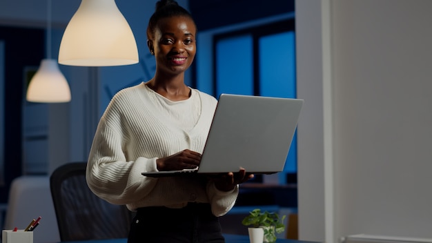 Mulher de negócios negra olhando para a frente sorrindo segurando um laptop em pé perto da mesa em uma empresa iniciante tarde da noite