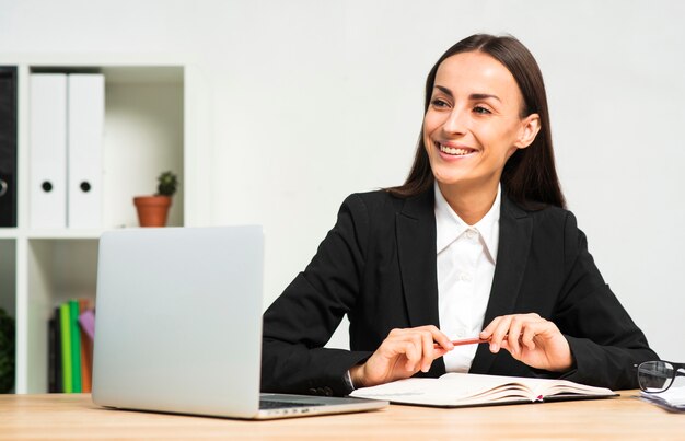 Mulher de negócios nova feliz que senta-se atrás da mesa com livro; caneta e laptop