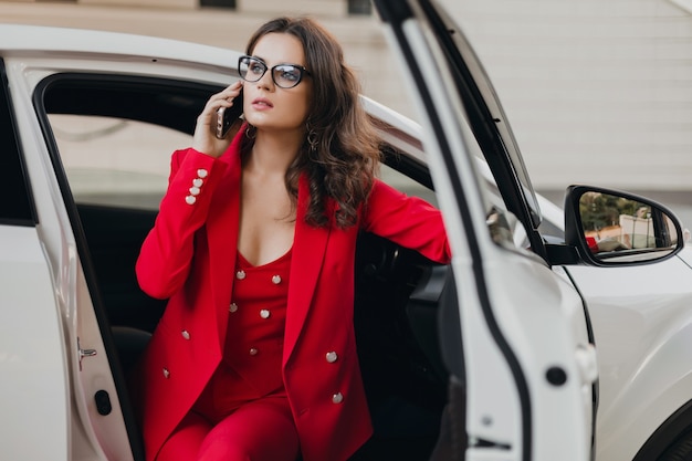 Mulher de negócios rica e sexy linda em um terno vermelho sentada em um carro branco, usando óculos, falando ao telefone, estilo de mulher de negócios