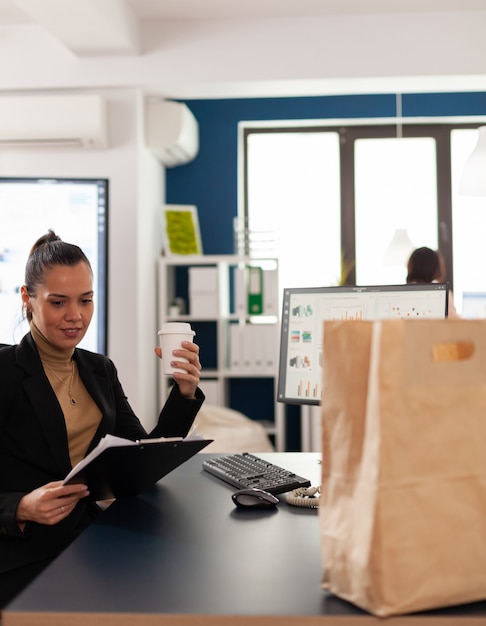 Mulher de negócios sentada à mesa no escritório corporativo lendo estatísticas financeiras na prancheta, antes de saborear a deliciosa comida para viagem em um saco de papel