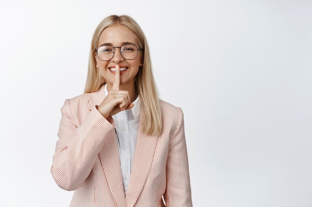 Mulher de negócios sorridente mostrando sinal de silêncio, mantendo um segredo, fazendo shh gesto, de terno e óculos em branco