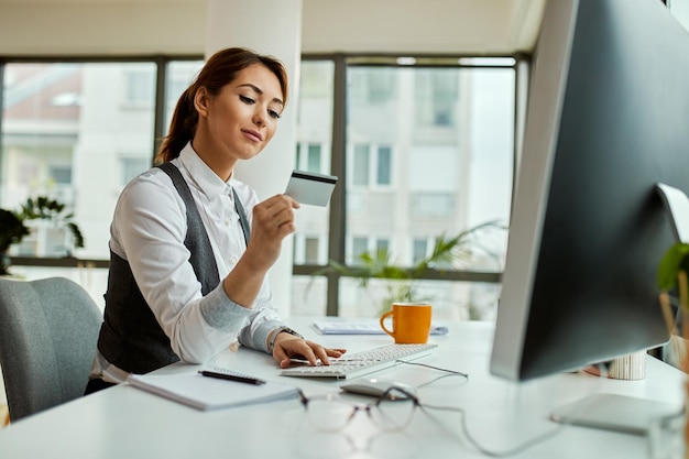 Mulher de negócios sorridente usando cartão de crédito e computador enquanto compra na internet no escritório