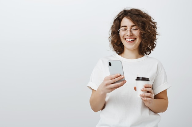 Mulher de óculos sorrindo para a mensagem de texto do celular, bebendo café no intervalo do escritório