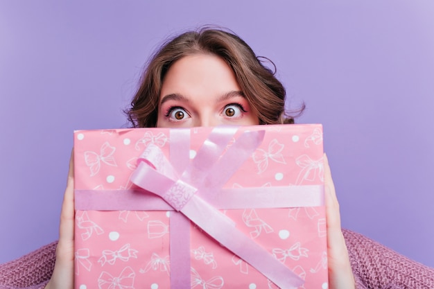 Foto grátis mulher de olhos castanhos chocada segurando um grande presente de aniversário na frente do rosto. retrato do close-up de uma menina morena espantada com presente de natal rosa em primeiro plano.