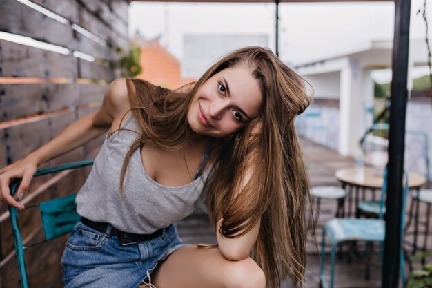 Mulher de olhos escuros em saia jeans brincando com os cabelos em um café ao ar livre. Modelo feminino muito moreno posando de manhã no restaurante de rua.