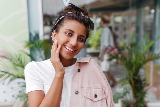 Mulher de olhos verdes em uma camiseta branca e jaqueta rosa com um sorriso branco como a neve olha para a frente, posando na rua