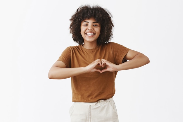 Foto grátis mulher de pele escura atraente positiva e feliz com penteado afro mostrando o sinal do coração sobre o peito e sorrindo com alegria e cuidado, tendo afeto ou expressando amor sobre uma parede cinza