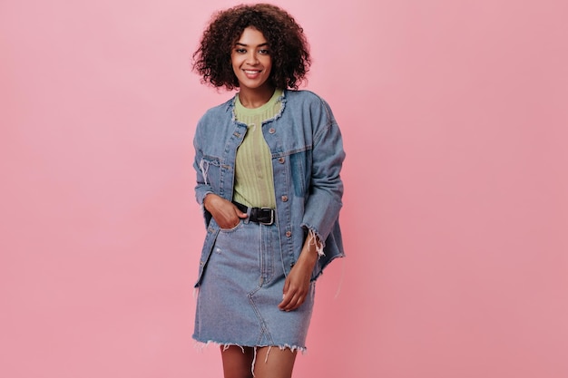 Mulher de pele escura com roupa jeans, olhando para a câmera no fundo rosa retrato de menina encaracolada em camiseta verde sorrindo em pano de fundo isolado