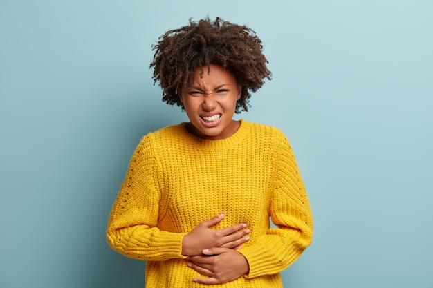 Mulher de pele escura descontente sofre de dor de estômago, cãibras menstruais, mantém as mãos na barriga, usa suéter de malha amarelo, sorri afetuosamente, fica de pé contra um fundo azul.