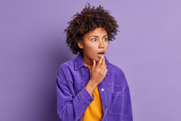 Mulher de pele escura emocional deixa cair o queixo e olha com espanto não espera ver algo medroso vestido com uma jaqueta brilhante.