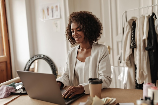 Mulher de pele escura encaracolada atraente trabalha no laptop e senta-se à mesa com uma xícara de café nela feliz designer posa no escritório aconchegante