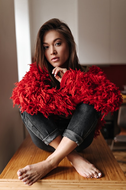 Mulher de pele escura está sentada na mesa com as pernas cruzadas. retrato de mulher de jaqueta de malha e jeans na cozinha.