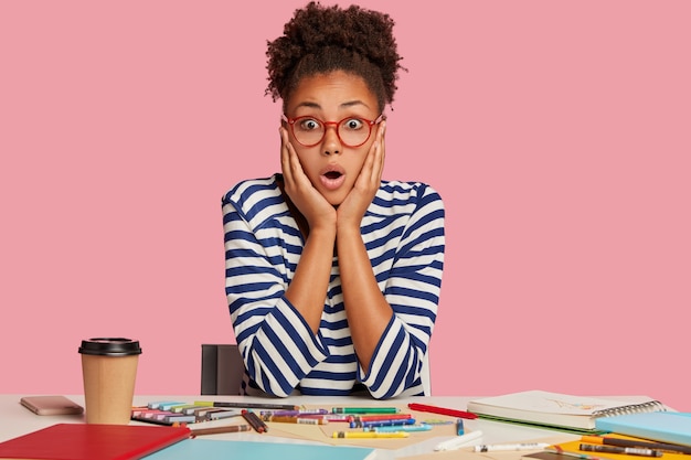 Foto grátis mulher de pele escura estupefata com expressão facial surpresa, mantém as mãos nas bochechas, olha aterrorizada, recebe muitas tarefas, posa no local de trabalho com giz de cera, diário em espiral, bebe café, usa óculos