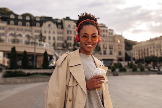 Foto grátis mulher de pele escura feliz segura copo de suco de laranja senhora encantadora de casaco bege ouve música em fones de ouvido vermelhos do lado de fora