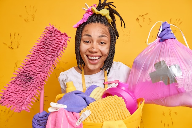 Foto grátis mulher de pele escura positiva fica suja após a limpeza geral fazer o trabalho doméstico segura o saco de lixo e o esfregão sujo arruma o apartamento lava a roupa sorrisos amplamente isolados sobre a parede amarela