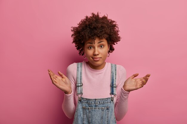 Mulher de pele escura, questionada e insegura, levanta as mãos com expressão confusa, intrigada para responder a uma pergunta difícil, usa roupa casual, isolada sobre parede rosa. Percepção humana