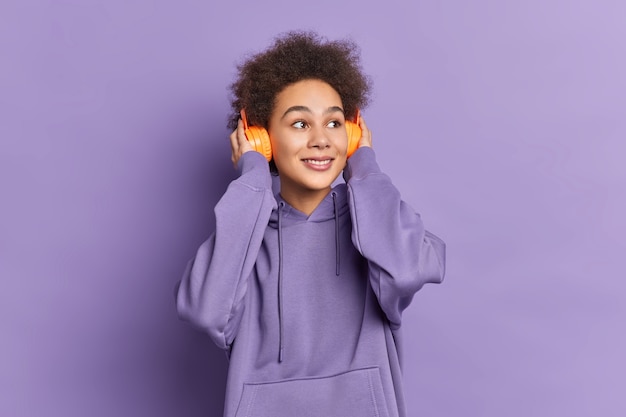 Mulher de pele escura satisfeita com cabelos crespos e encaracolados usa fones de ouvido estéreo e escuta música vestida de moletom.