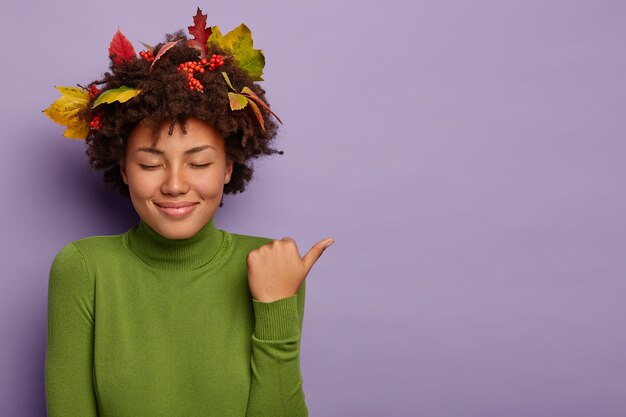 Mulher de pele escura satisfeita fecha os olhos de prazer, sorri gentilmente, aponta o polegar para longe, usa gola olímpica verde, demonstra promoção incrível