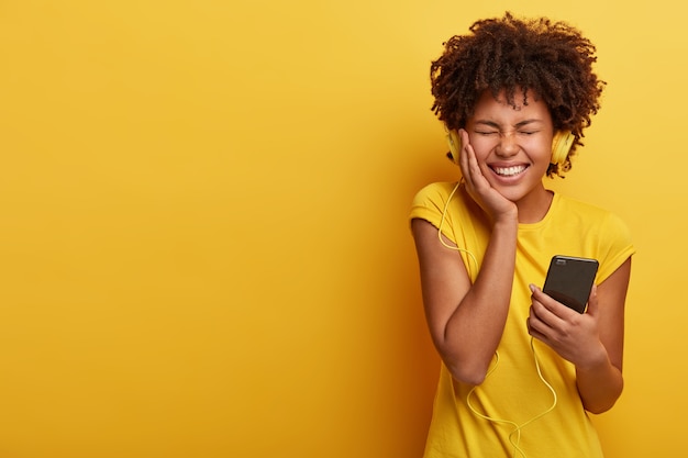 Foto grátis mulher de pele escura satisfeita se sente bem e feliz, sorri amplamente, segura o smartphone conectado a fones de ouvido, fecha os olhos de prazer
