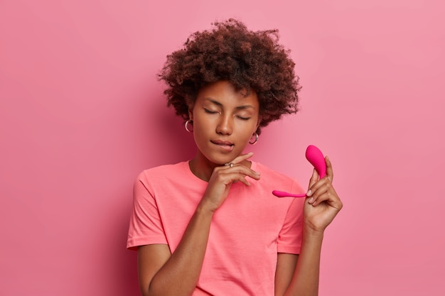 Mulher de pele escura satisfeita segura o brinquedo sexual do vibrador com app para controlar as vibrações, morde os lábios e fecha os olhos com satisfação, segura o ovo do vibrador inteligente de silicone isolado na parede rosa. prazer erótico