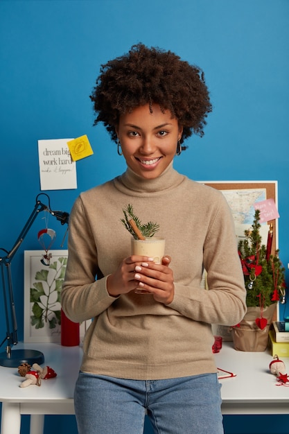 Foto grátis mulher de pele escura satisfeita segurando coquetel caseiro, vestida casualmente, com expressão alegre no rosto, aprecia o inverno e gemada saborosa inclinada na mesa branca com pequena árvore de natal