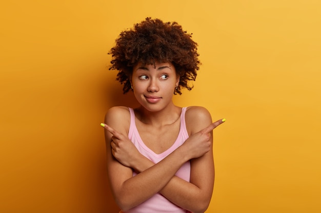 Mulher de pele morena confusa e duvidosa aponta para o lado, mantém as mãos cruzadas sobre o corpo, tenta decidir o que melhor escolher, vestida com camisa casual, não tem ideia, faz escolha, decide o que comprar