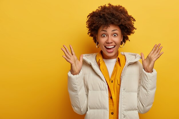 Mulher de pele morena surpresa e alegre levanta as palmas das mãos, feliz por receber um bom presente, usa um casaco branco quente de inverno, jaqueta amarela, expressa boas emoções, sorri amplamente, tem uma reação feliz na surpresa