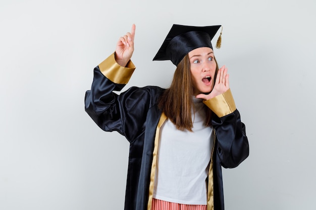 Mulher de pós-graduação apontando para cima, mantendo a mão perto da boca aberta em roupas casuais, uniforme e parecendo espantada. vista frontal.