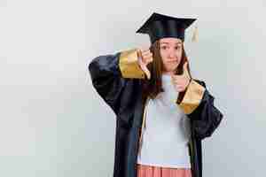 Foto grátis mulher de pós-graduação mostrando os polegares para cima e para baixo em roupas casuais, uniforme e parecendo indecisa. vista frontal.