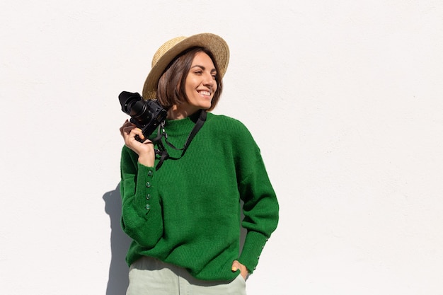 Mulher de suéter casual verde e chapéu ao ar livre na parede branca turista feliz positiva com câmera profissional