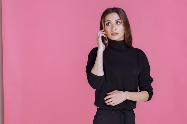 Mulher de suéter preto segurando um smartphone na orelha e falando.