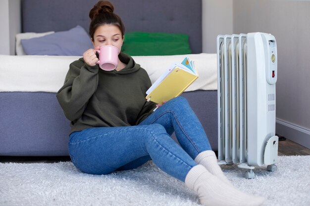 Mulher de tiro completo lendo livro perto de aquecedor